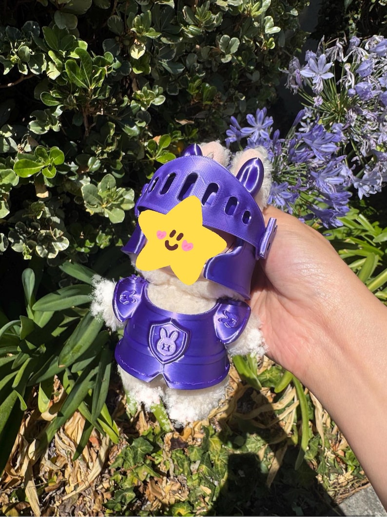 Op de afbeelding: Een wit pluche konijn met een paarse ridderhelm en -pantser. Het pantser heeft een konijnenwapen. Het konijn wordt vastgehouden voor een achtergrond van groen gebladerte en paarse bloemen.