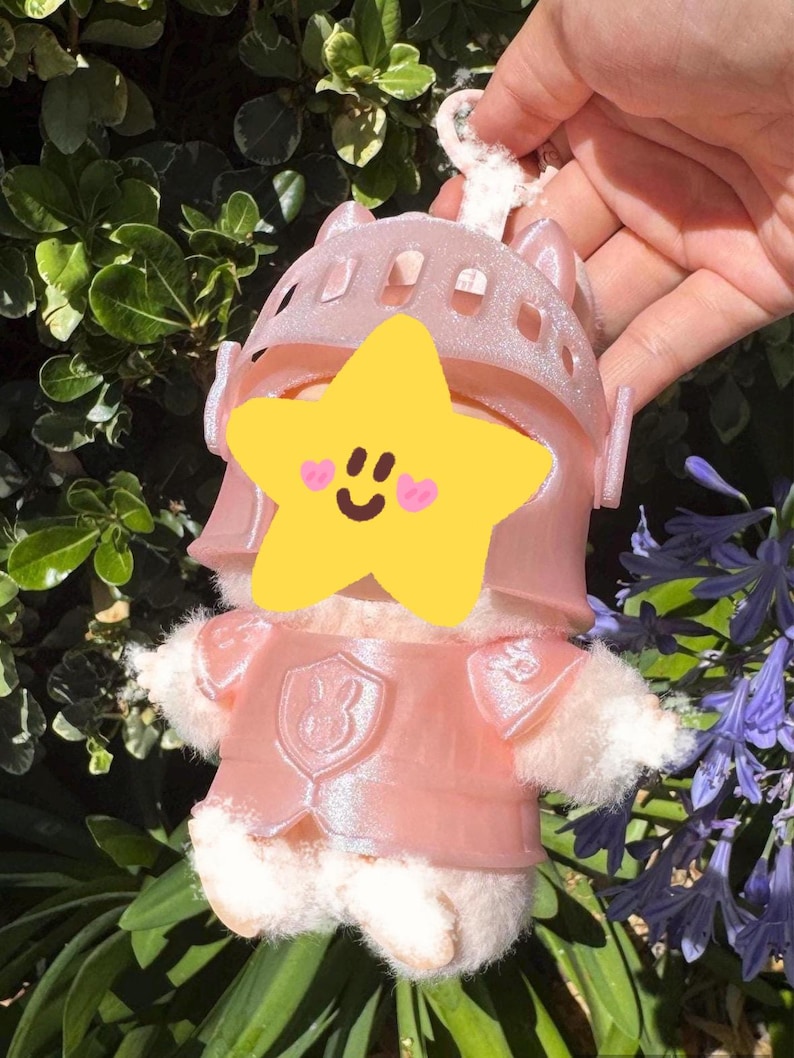 Op de afbeelding: Een roze ridderknuffel met helm en harnas. Het speelgoed heeft een pluizig wit lichaam en een stervormig gezicht. Het wordt vastgehouden door een wit touwtje.