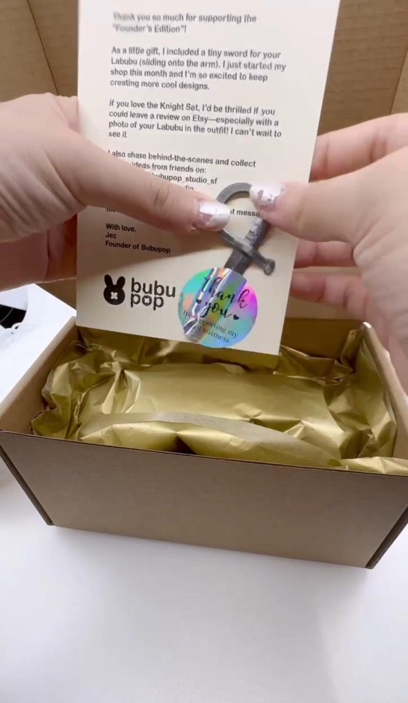 May include: A small cardboard box with gold tissue paper inside. A card with text and a small metal sword charm are visible. The card has a holographic sticker that says "Thank you". The box is being opened.