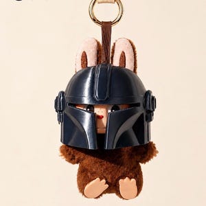 May include: A plush toy keychain featuring a brown furry body and a dark blue helmet. The toy has tan bunny ears and a small face peeking out from under the helmet. A gold ring and a tan strap are attached to the top.
