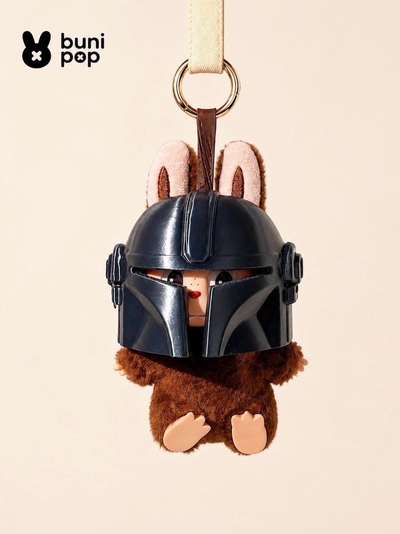 May include: A plush toy keychain featuring a brown furry body and a dark blue helmet. The toy has tan bunny ears and a small face peeking out from under the helmet. A gold ring and a tan strap are attached to the top.
