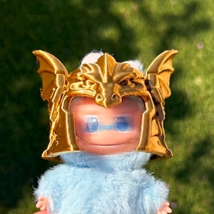 May include: A small figurine featuring a light blue fuzzy body and a shiny gold-colored helmet. The helmet is molded with dragon-like wings on the sides and a central crest. The face is smooth and light pink with two large, round blue eyes.