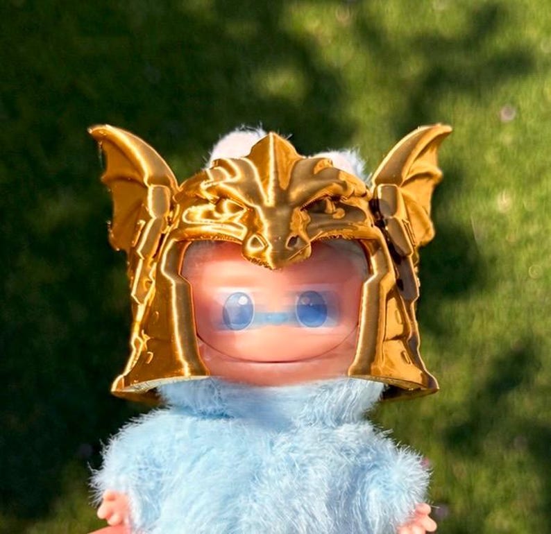May include: A small figurine featuring a light blue fuzzy body and a shiny gold-colored helmet. The helmet is molded with dragon-like wings on the sides and a central crest. The face is smooth and light pink with two large, round blue eyes.
