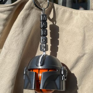 Puede incluir: Un llavero con forma de casco mandaloriano plateado con la palabra "MANDO" escrita con cuentas cuadradas. El casco tiene un dise&ntilde;o de baloncesto naranja y negro.