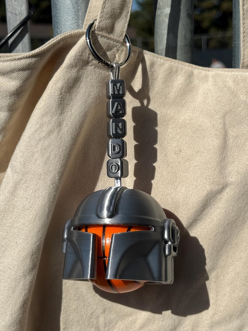 Puede incluir: Un llavero con forma de casco mandaloriano plateado con la palabra "MANDO" escrita con cuentas cuadradas. El casco tiene un dise&ntilde;o de baloncesto naranja y negro.