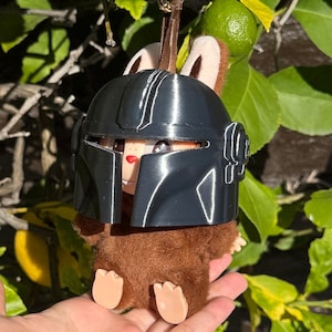 May include: A brown plush toy with a black helmet, brown ears, and tan feet. The toy is hanging from a gold-colored ring and brown strap. The background includes green leaves and a lime.