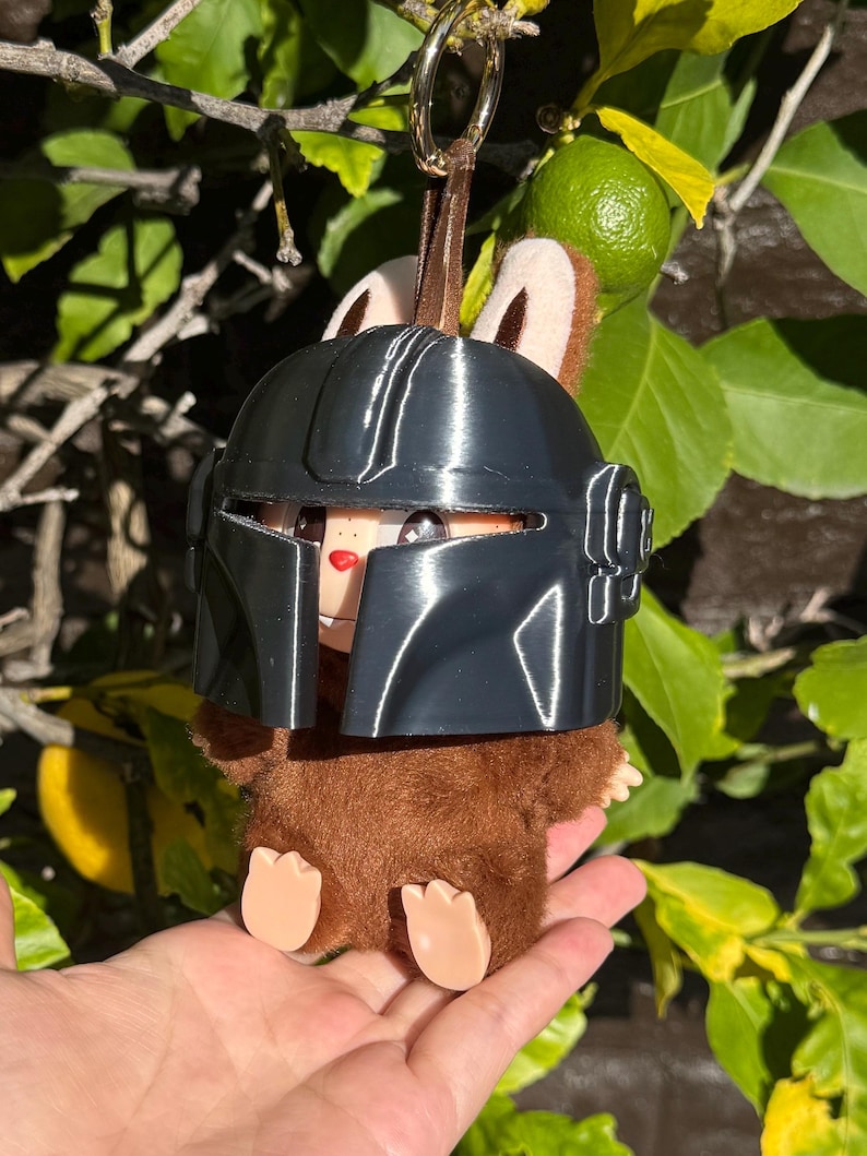 May include: A brown plush toy with a black helmet, brown ears, and tan feet. The toy is hanging from a gold-colored ring and brown strap. The background includes green leaves and a lime.