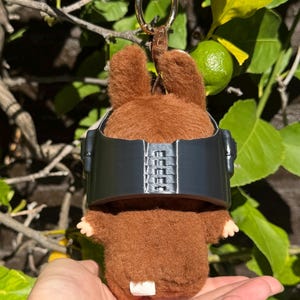 May include: A brown plush toy bunny with a black visor and a gold keychain. The bunny has small ears and tiny hands. The background features green leaves and a lime.