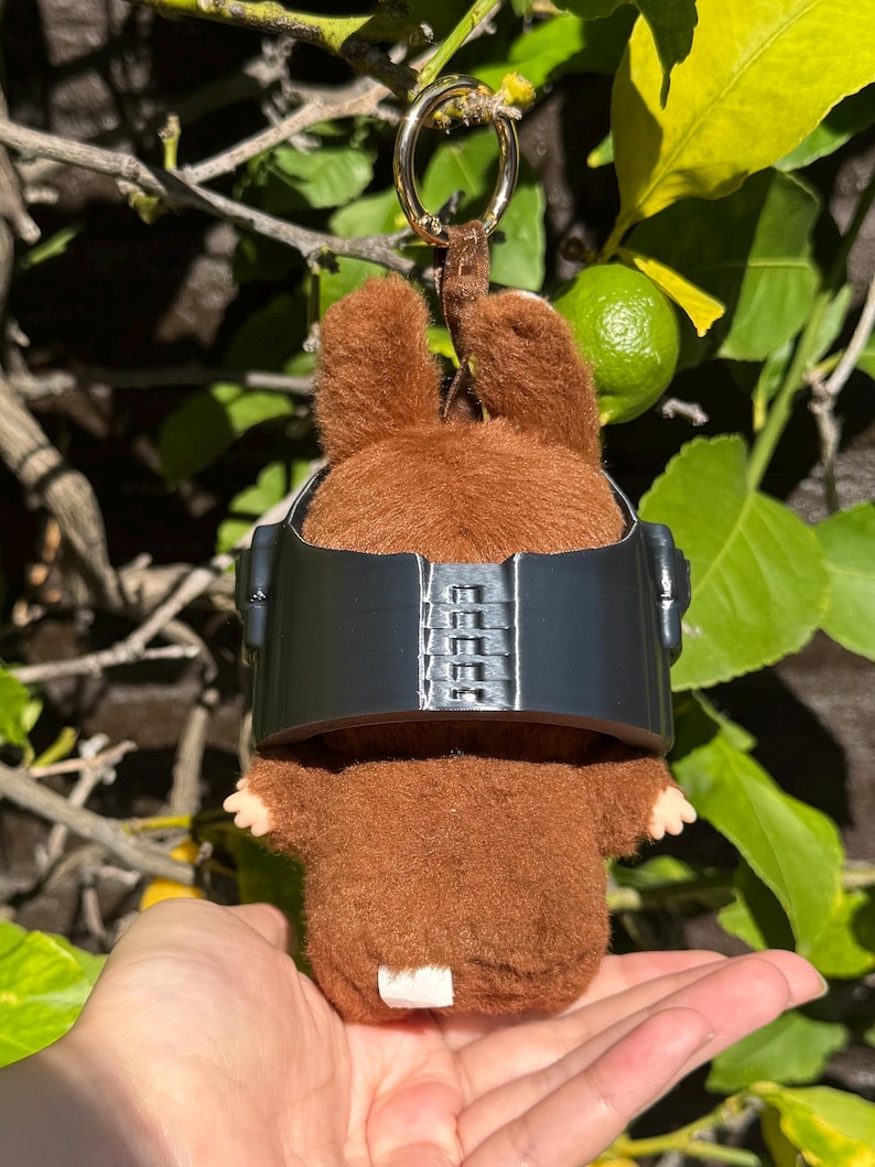 May include: A brown plush toy bunny with a black visor and a gold keychain. The bunny has small ears and tiny hands. The background features green leaves and a lime.