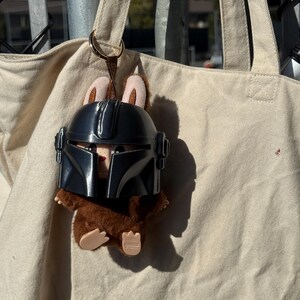 May include: A plush toy featuring a brown creature wearing a black Mandalorian helmet. The toy has tan ears and is attached to a beige tote bag with a gold-colored ring.