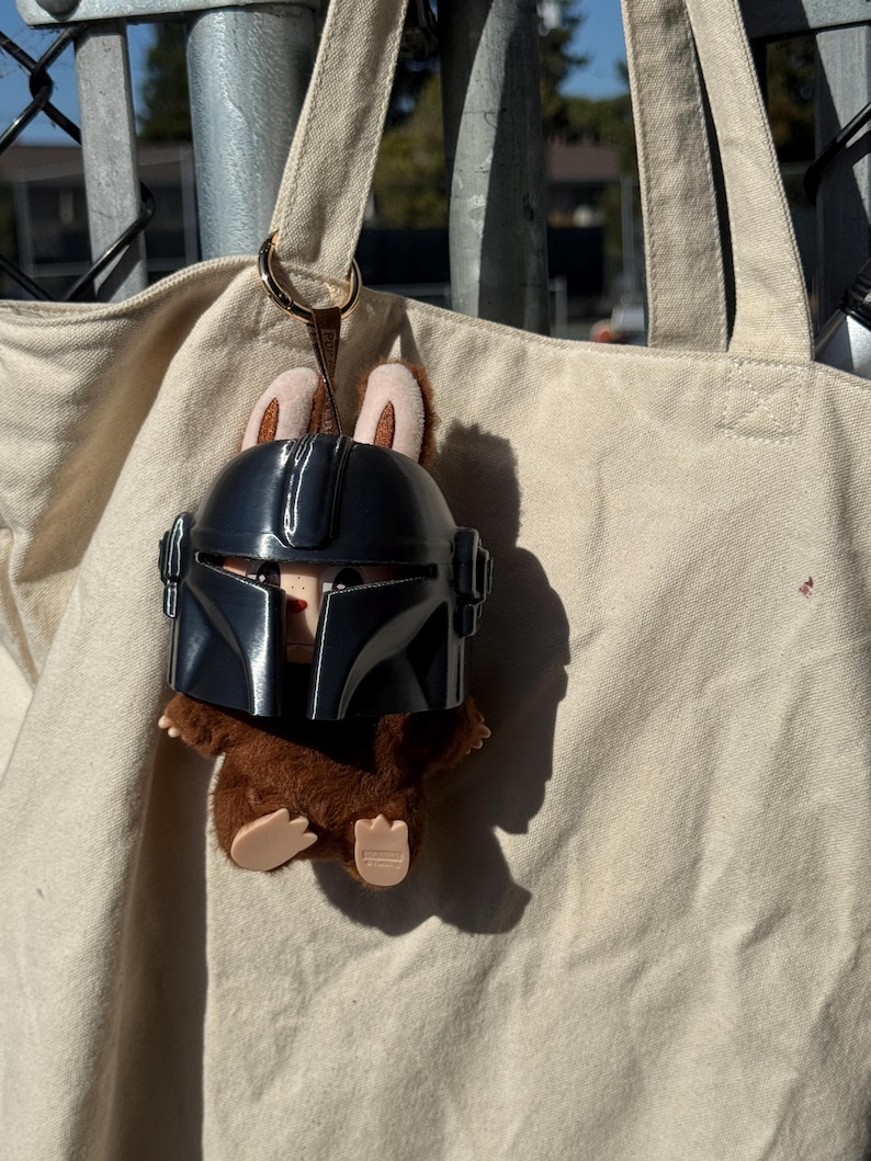May include: A plush toy featuring a brown creature wearing a black Mandalorian helmet. The toy has tan ears and is attached to a beige tote bag with a gold-colored ring.