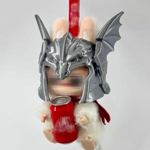 May include: A plush white rabbit figure wearing a metallic silver dragon-shaped helmet with wings. The figure has a red cape and is holding a small red cylindrical can. A red plastic loop is attached to the top of the helmet for hanging.