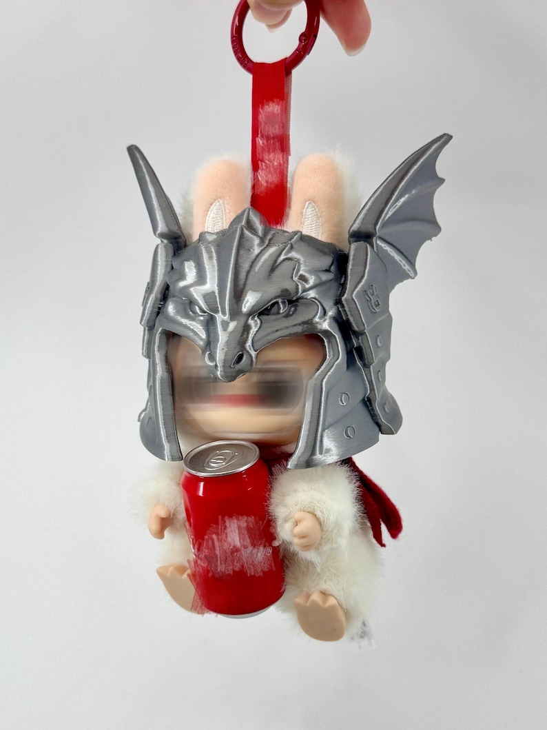 May include: A plush white rabbit figure wearing a metallic silver dragon-shaped helmet with wings. The figure has a red cape and is holding a small red cylindrical can. A red plastic loop is attached to the top of the helmet for hanging.