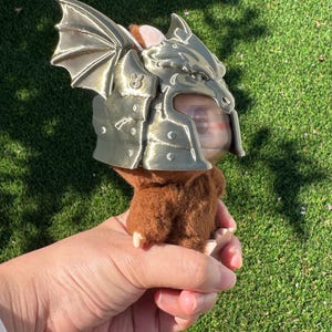 May include: A small brown plush creature wearing a metallic gold-colored dragon helmet. The helmet features a sculpted dragon head with prominent wings on the side and a small rabbit icon embossed on the temple area. The plush has soft brown fur.