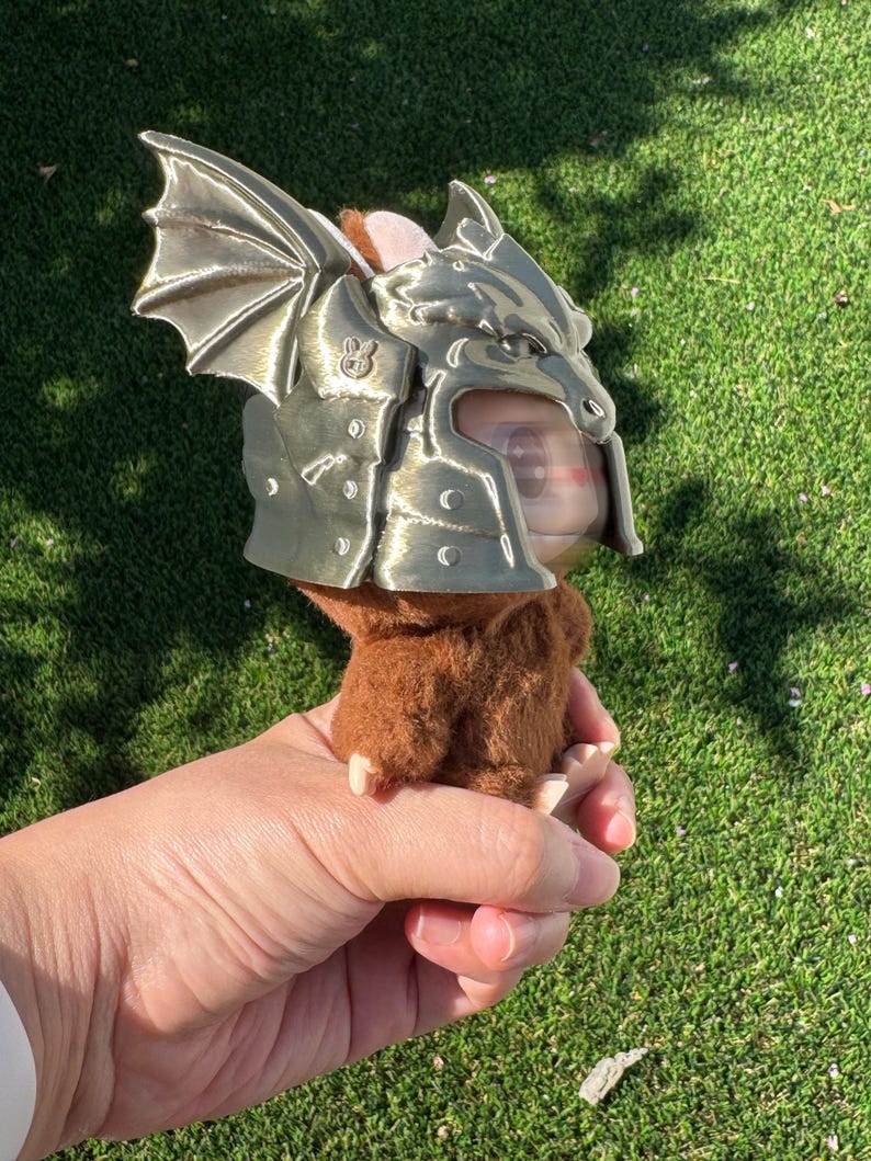 May include: A small brown plush creature wearing a metallic gold-colored dragon helmet. The helmet features a sculpted dragon head with prominent wings on the side and a small rabbit icon embossed on the temple area. The plush has soft brown fur.
