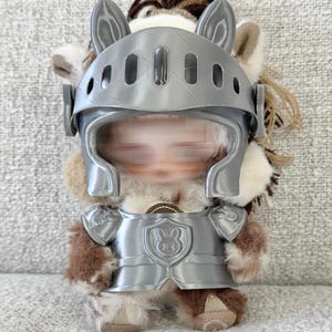 Może przedstawiać: Pluszowa zabawka ubrana w srebrny hełm i zbroję rycerską. Hełm ma detale w kształcie uszu, a zbroja tarczę z emblematem królika. Zabawka ma brązowe i beżowe futro.