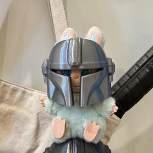 May include: A plush toy bunny wearing a silver Mandalorian helmet. The bunny has light blue fur, pink ears, and pink paws. The helmet is detailed with ridges and a visor. The toy is set against a neutral background.
