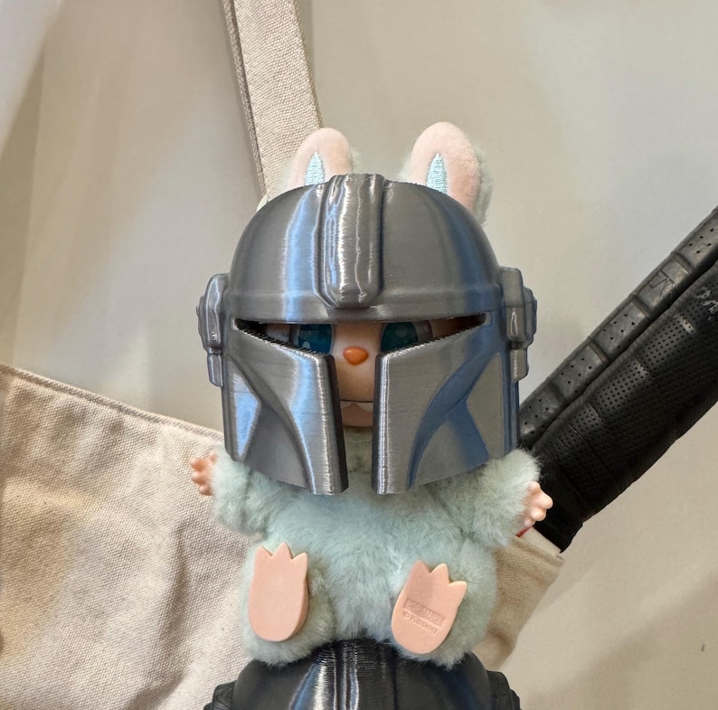 May include: A plush toy bunny wearing a silver Mandalorian helmet. The bunny has light blue fur, pink ears, and pink paws. The helmet is detailed with ridges and a visor. The toy is set against a neutral background.
