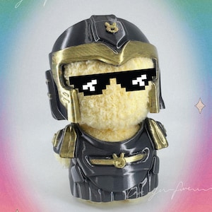 May include: A plush toy wearing a black and gold helmet and armor, with pixelated sunglasses. The toy has a light yellow body and is set against a colorful, blurred background. The words "love you forever" are written in cursive.