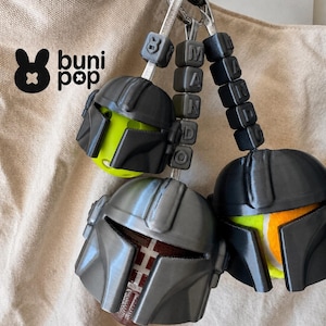 Puede incluir: Tres llaveros con dise&ntilde;os de casco y la palabra "MANDO" deletreada en cuentas de letras negras. Los cascos son grises y negros, con acentos verde ne&oacute;n, rojos y amarillos. El logotipo "buni pop" es visible en la esquina superior izquierda.