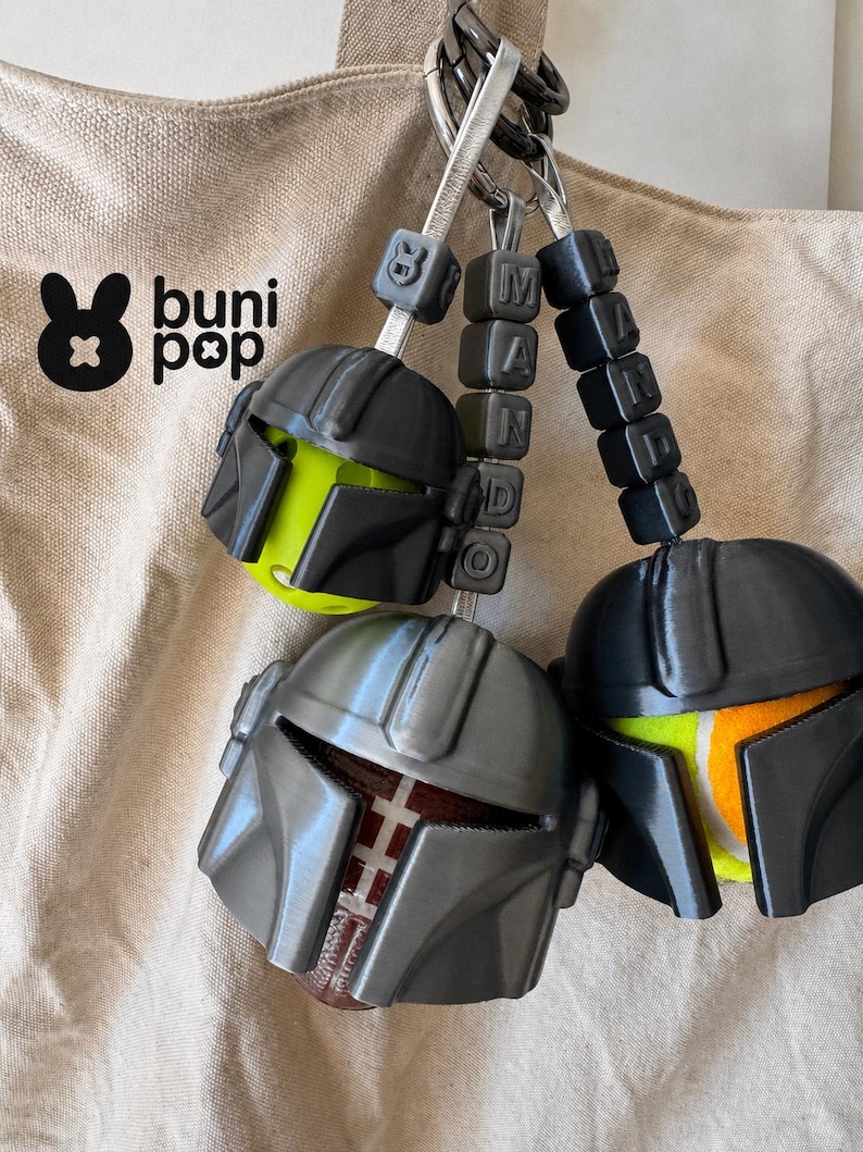 Puede incluir: Tres llaveros con dise&ntilde;os de casco y la palabra "MANDO" deletreada en cuentas de letras negras. Los cascos son grises y negros, con acentos verde ne&oacute;n, rojos y amarillos. El logotipo "buni pop" es visible en la esquina superior izquierda.