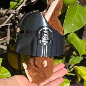 May include: A plush toy with brown fur and a black helmet. The toy is hanging from a brown ribbon. The helmet has a detailed design with a symbol on the front. The background features green leaves and a lime.