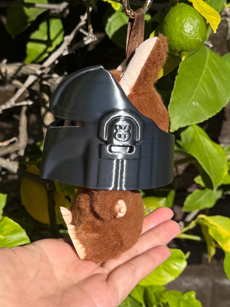 May include: A plush toy with brown fur and a black helmet. The toy is hanging from a brown ribbon. The helmet has a detailed design with a symbol on the front. The background features green leaves and a lime.