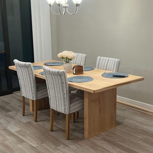 May include: A light wood dining table with a single pedestal base and four chairs. The chairs have blue and white striped fabric upholstery. The table is set with placemats and a vase of white flowers.