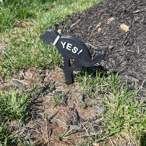 May include: A black, dog-shaped lawn stake with a white collar and the word YES! printed in white on its side. The silhouette depicts a dog in a squatting position, designed to be inserted into the ground as a marker for pet waste areas.