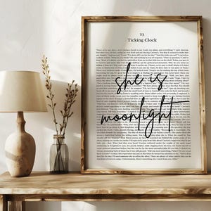 May include: A framed print with the words "she is moonlight" in a handwritten font, set against a backdrop of book pages. The frame is a light brown color. A wooden lamp and a vase with dried flowers are on a wooden table.