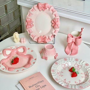 May include: A set of pink ceramic tableware with bow motifs. The collection includes plates, a cup with a spoon, and a small vase. Some plates are decorated with strawberries. A book titled "Butterflies On Your Lips" is also visible.