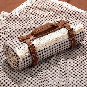 May include: Rolled picnic blanket with a brown and white checkered pattern, secured with brown leather straps and a handle. The blanket features a cream-colored lace trim, creating a rustic and stylish look. The leather straps have gold-colored buckles.