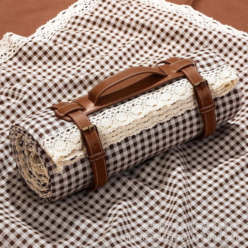 May include: Rolled picnic blanket with a brown and white checkered pattern, secured with brown leather straps and a handle. The blanket features a cream-colored lace trim, creating a rustic and stylish look. The leather straps have gold-colored buckles.