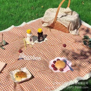 May include: A picnic scene with a red and white checkered blanket, a wicker basket, and food items. The blanket has a white lace trim. There are two decorative condiment bottles, a plate with grapes and a bun, and a small container with a sandwich.