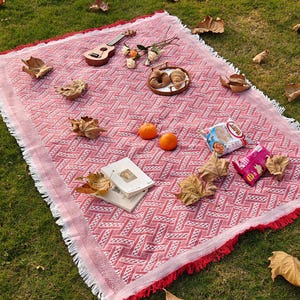 Op de afbeelding: Een rood-wit patroon picknickdeken met een gefranjerde rand, uitgespreid op groen gras. De deken is versierd met snacks, boeken, sinaasappels en een ukelele. Gedroogde bladeren en bloemen dragen bij aan de buitenscène.