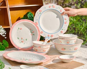 May include: A collection of white ceramic tableware with a strawberry theme. The set includes plates, bowls, and spoons, all featuring pink and blue accents. The plates and bowls have a scalloped edge design.