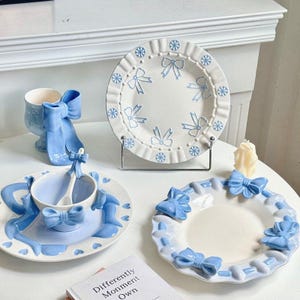 May include: A collection of light blue and white ceramic tableware featuring bow and snowflake designs. The set includes plates, a teacup with a saucer and spoon, and a decorative cup. The pieces are arranged on a white surface.