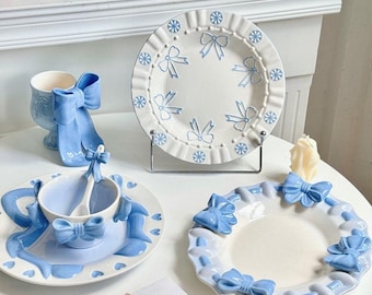 May include: A collection of light blue and white ceramic tableware featuring bow and snowflake designs. The set includes plates, a teacup with a saucer and spoon, and a decorative cup. The pieces are arranged on a white surface.
