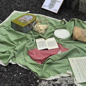 May include: A picnic scene with a green and white checkered blanket with a white lace trim. A red and white checkered napkin, an open book, a basket, a plate, and a decorative box are on the blanket. A small bag and a sign are also present.