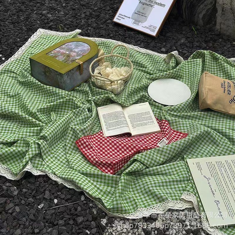 May include: A picnic scene with a green and white checkered blanket with a white lace trim. A red and white checkered napkin, an open book, a basket, a plate, and a decorative box are on the blanket. A small bag and a sign are also present.