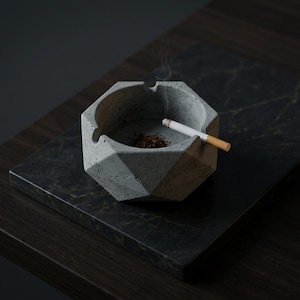 Peut inclure: Un cendrier géométrique en béton gris avec une cigarette allumée et du tabac repose sur un plateau en marbre foncé. Le cendrier a un design à facettes. De la fumée s'élève de la cigarette.