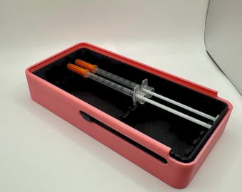May include: A pink rectangular case with a black interior holds two syringes with orange caps. The case has a sliding mechanism on the side. The syringes are clear with white plungers.