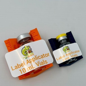 May include: Two small glass vials with silver and yellow caps, each labeled with "Label Applicator" and the vial size. One vial is in an orange holder, the other in a blue holder. The labels feature a green chameleon logo.