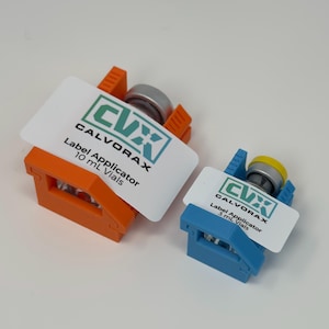 May include: Two label applicators, one orange and one blue, each holding a vial. The applicators have white labels with the text "CVX CALVORAX Label Applicator" and vial size information. The vials have silver and yellow caps.
