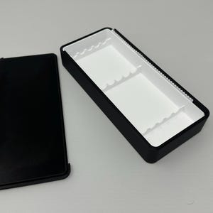 May include: A black rectangular box with a white interior and a separate black lid. The box has a rectangular shape with a black exterior and a white interior with dividers. The lid is also black and rectangular.