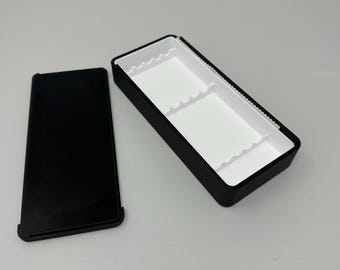 May include: A black rectangular box with a white interior and a separate black lid. The box has a rectangular shape with a black exterior and a white interior with dividers. The lid is also black and rectangular.