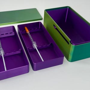 May include: A set of purple and green storage containers with a lid. The containers have a rectangular shape and are designed to hold medical syringes. The syringes have orange caps.