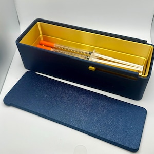 May include: A navy blue rectangular storage box with a gold interior and a matching lid. Inside the box are two syringes with orange plungers. The box is open, revealing the contents. The lid is placed in front of the box.