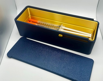 May include: A navy blue rectangular storage box with a gold interior and a matching lid. Inside are two syringes with orange plungers. The box is designed to hold medical supplies. The lid is placed next to the box.