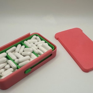 May include: A pink pill organizer with a green interior and a separate pink lid. The organizer is filled with white oval pills. The design includes a sliding mechanism and a green button for opening and closing.